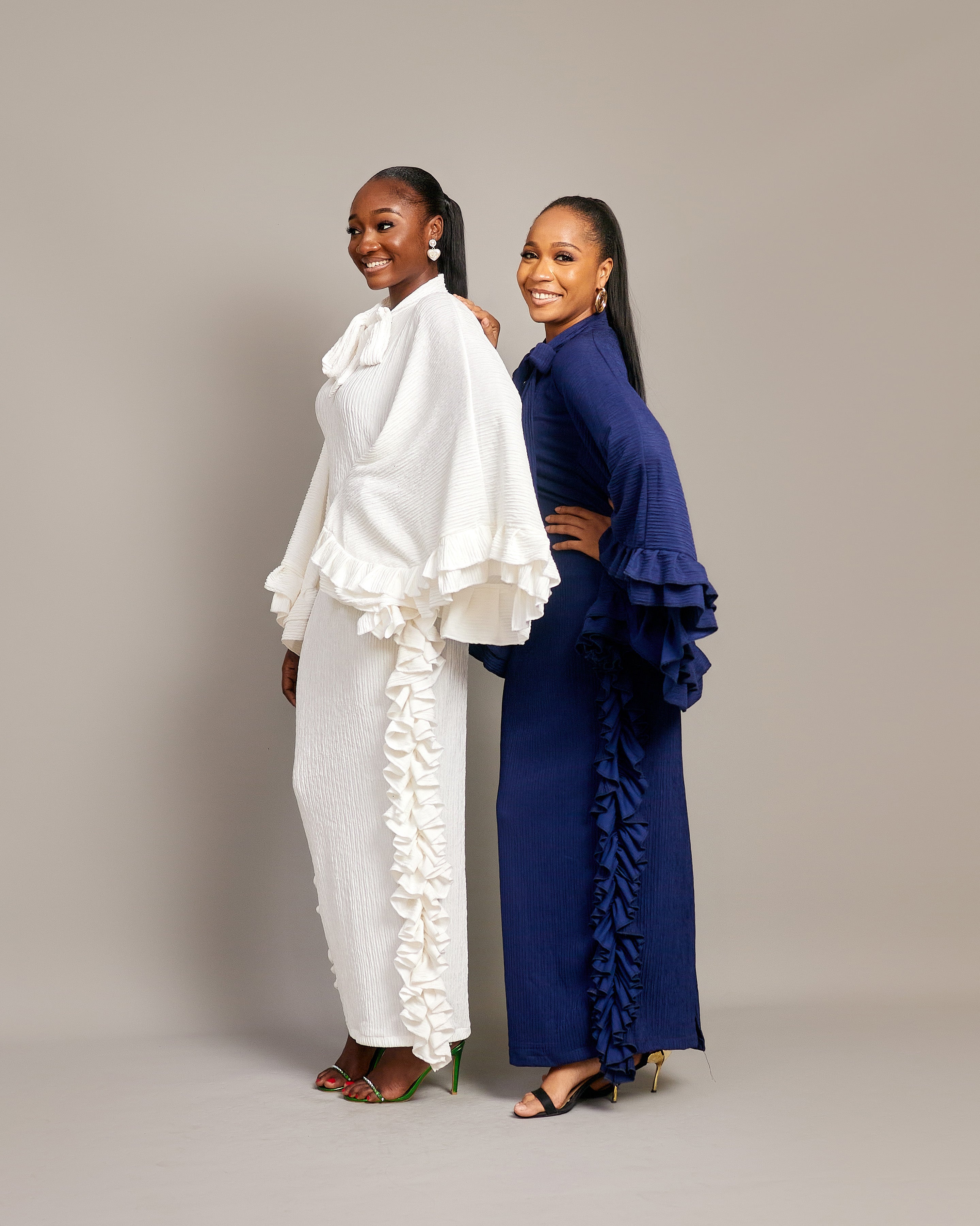 Two women wearing the Tella Dress: one in ivory, one in navy. Maxi gowns with ruffle sides, long sleeves, and bow-tie neckline.

