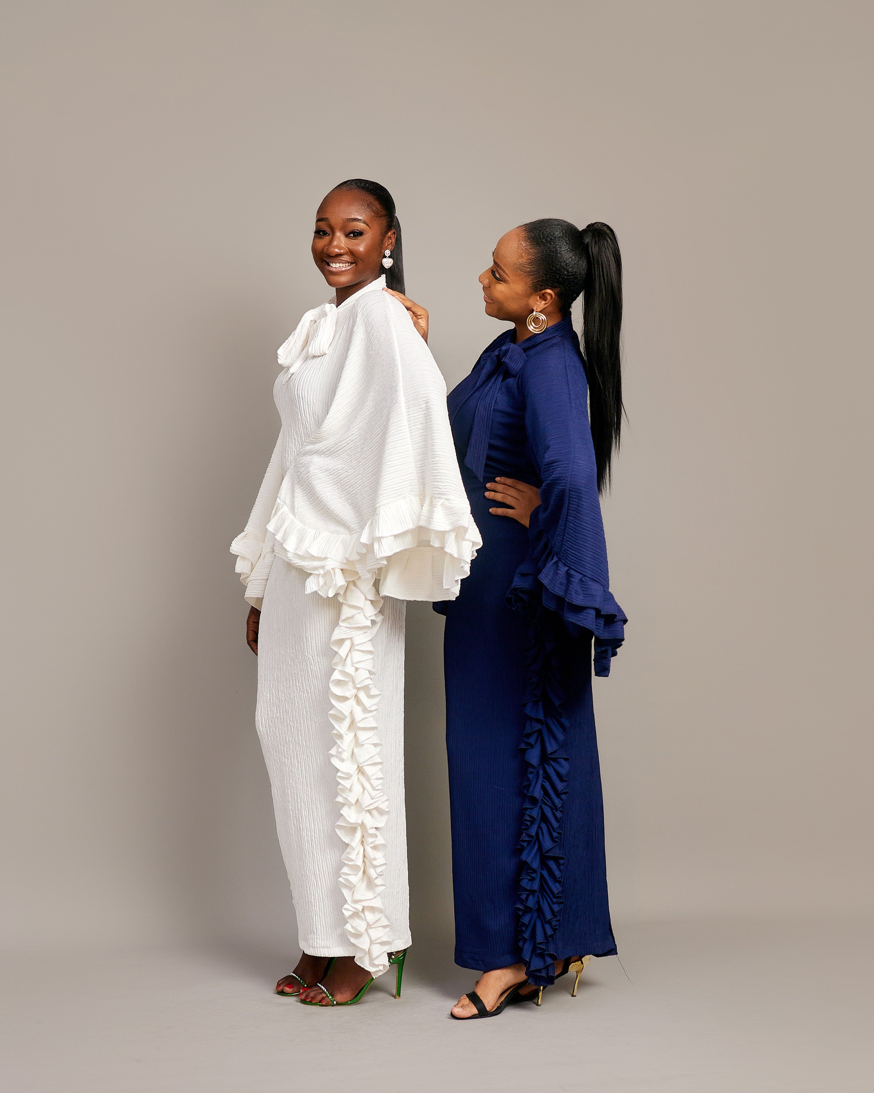 Two women wearing the Tella Dress: one in ivory, one in navy. Maxi gowns with ruffle sides, long sleeves, and bow-tie neckline.
