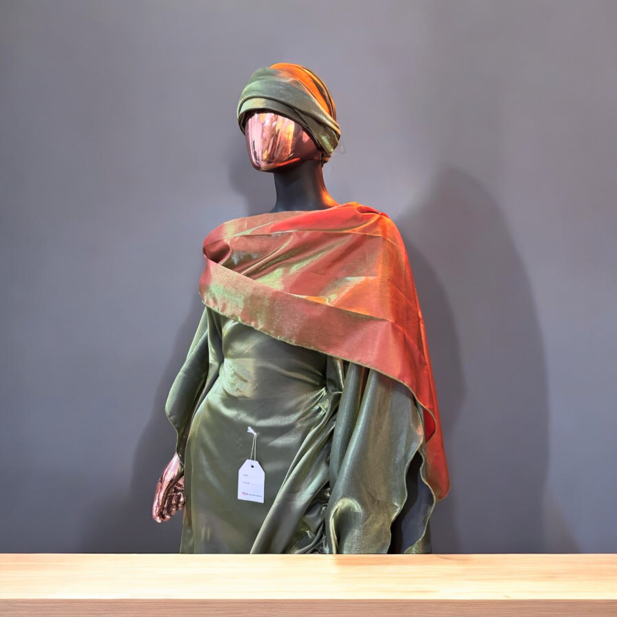 Olive green satin gown with ruched side draping, paired with a bronze-toned shawl and matching headwrap, displayed on a mannequin in an arched interior.
