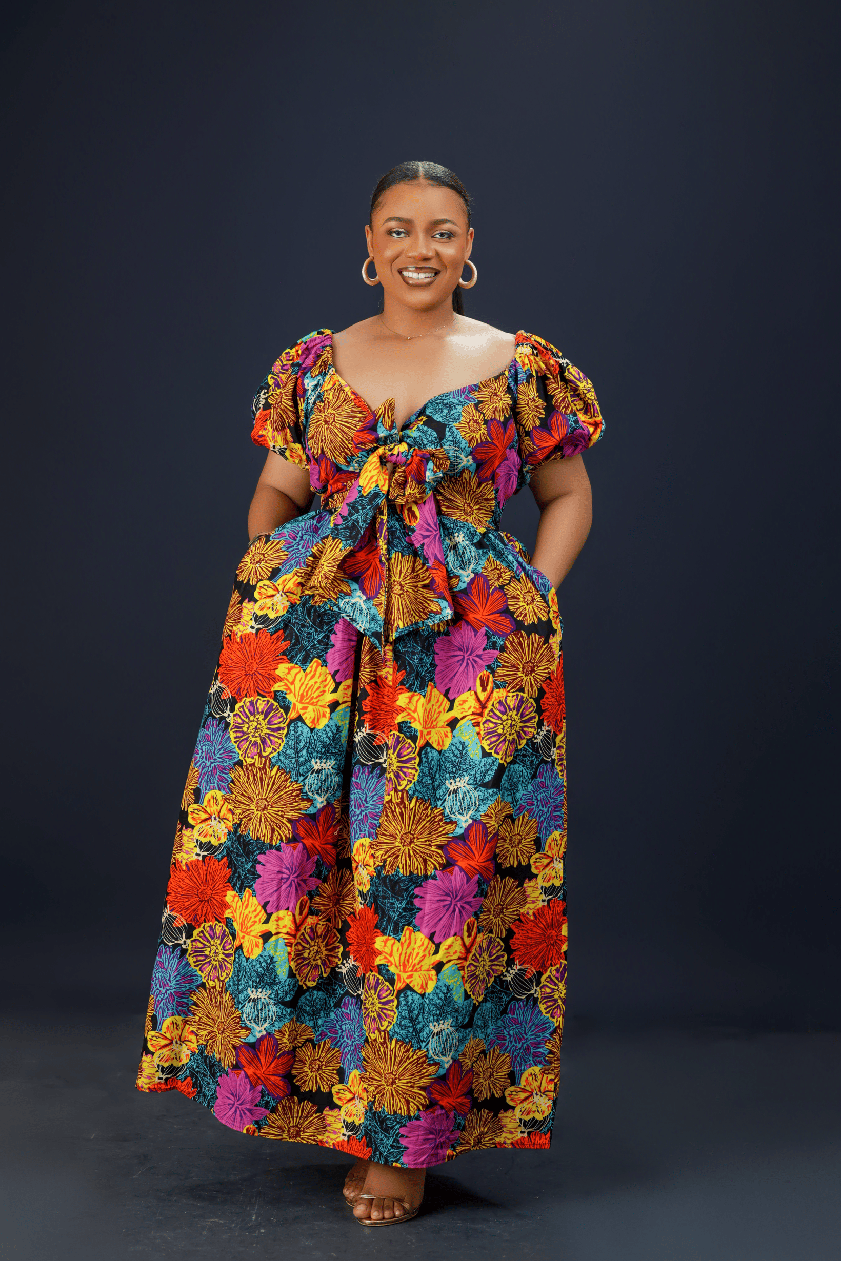 A woman wearing a vibrant floral maxi dress with puff sleeves, a front bow detail, and side pockets against a dark background.
