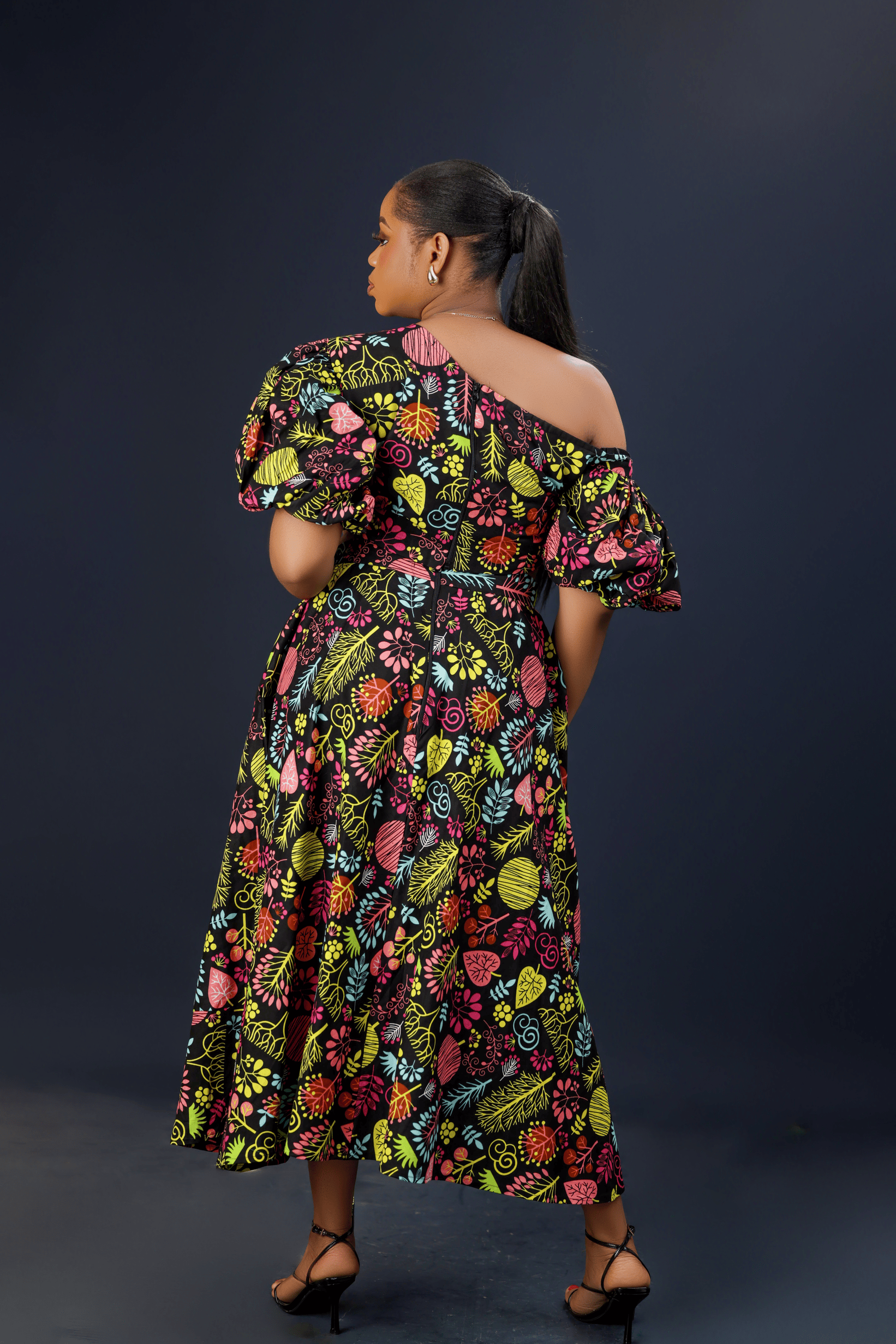 A woman wearing a vibrant floral maxi dress with puff sleeves, a front bow detail, and side pockets against a dark background.
