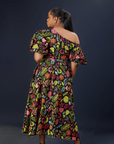A woman wearing a vibrant floral maxi dress with puff sleeves, a front bow detail, and side pockets against a dark background.
