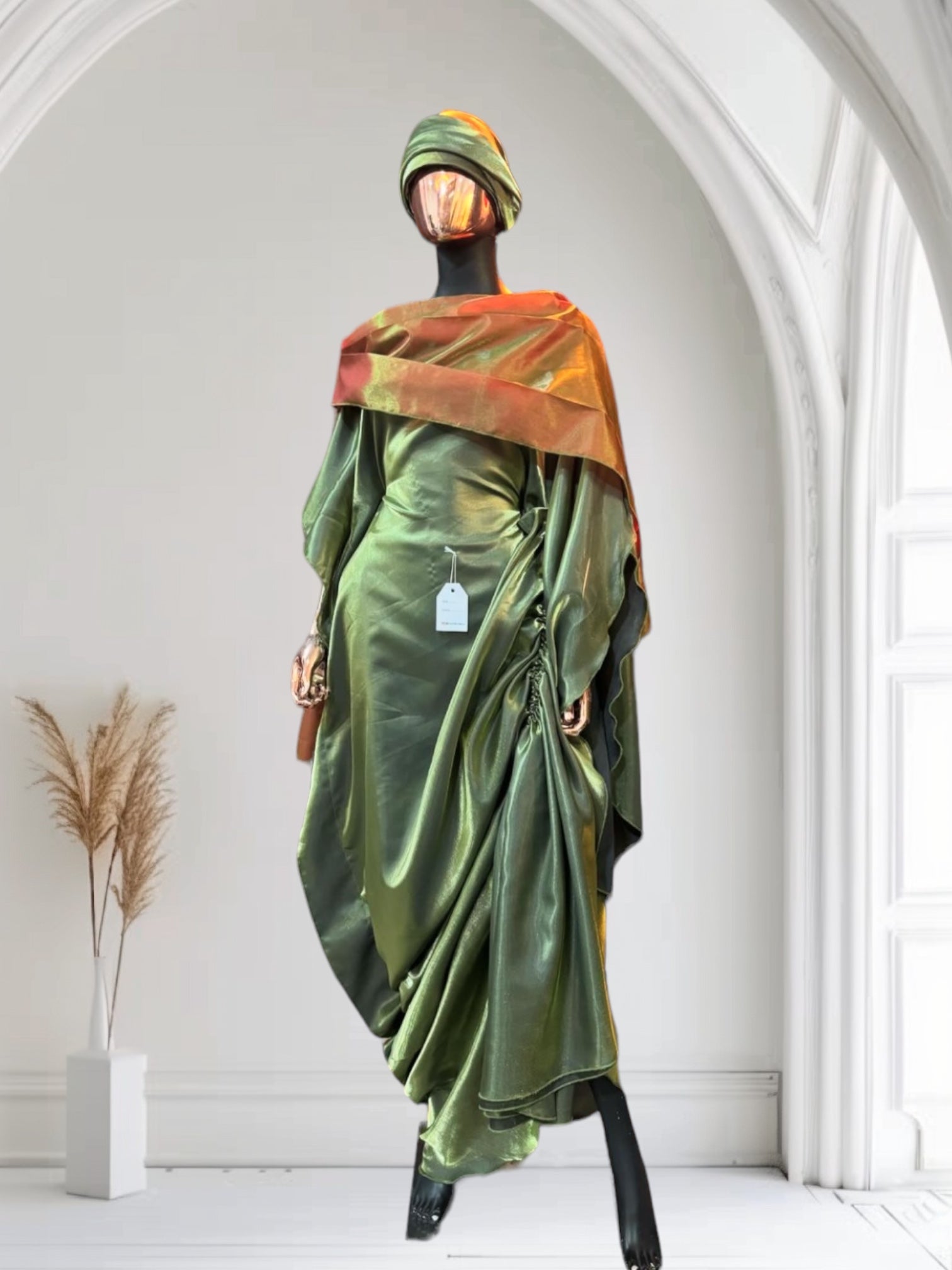 Olive green satin gown with ruched side draping, paired with a bronze-toned shawl and matching headwrap, displayed on a mannequin in an arched interior.
