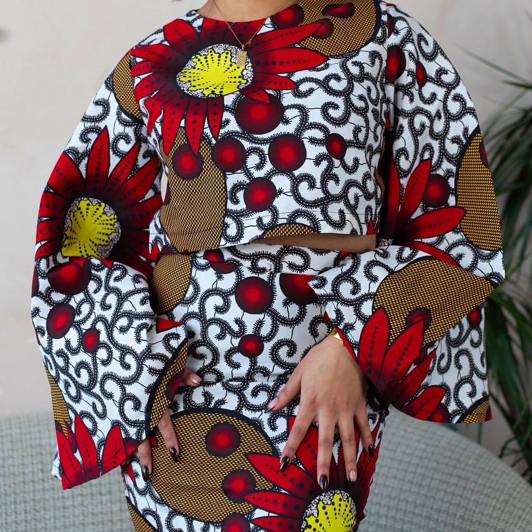 Woman wearing a bold Ankara two-piece set with flared sleeves and a fitted skirt in red, yellow, and black floral prints.
