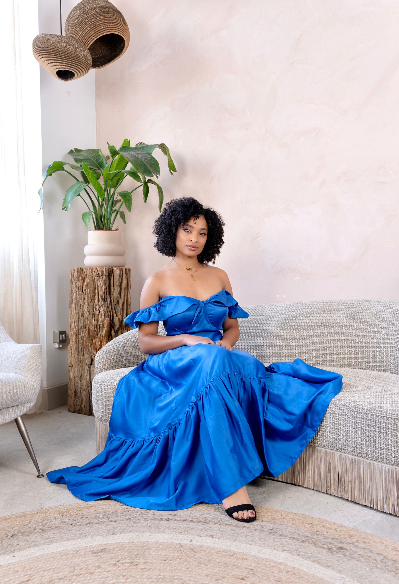 Model wearing a royal blue satin off-shoulder crop top and matching flared skirt in a minimalist living room setting.
